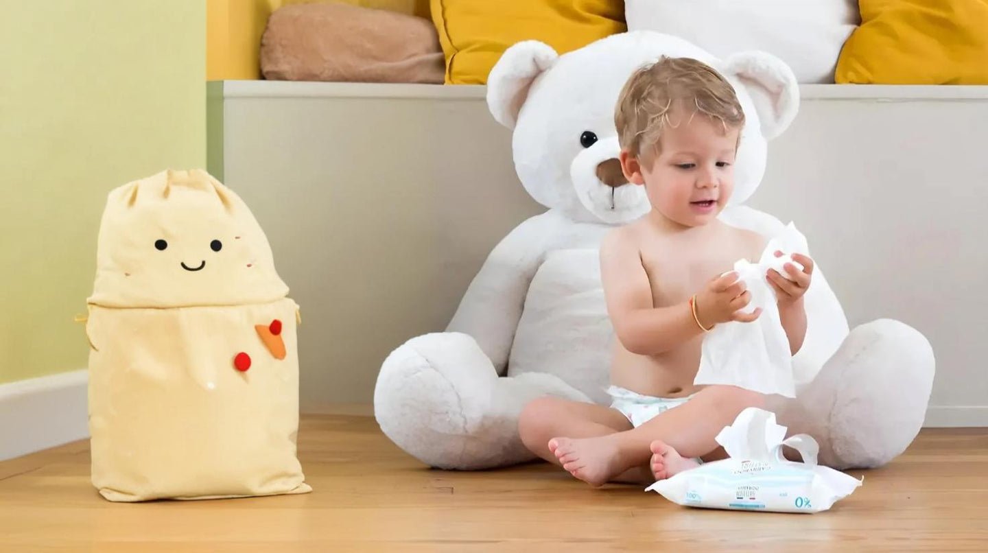 Enfant assis sur le sol avec un grand ours en peluche, jouant avec des lingettes, à côté d'un sac de rangement souriant