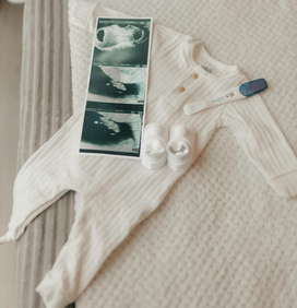 Body blanc avec petits chaussons bébé dessus, un test de grossesse positif et une écographie de bébé, sur un drap de lit blanc 