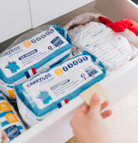 Main qui ouvre un placard de rangement où on aperçoit des paquets de couches taille 1 Carryboo de nos Régions, ainsi que des petites couches Carryboo de nos Régions dans un pochon 