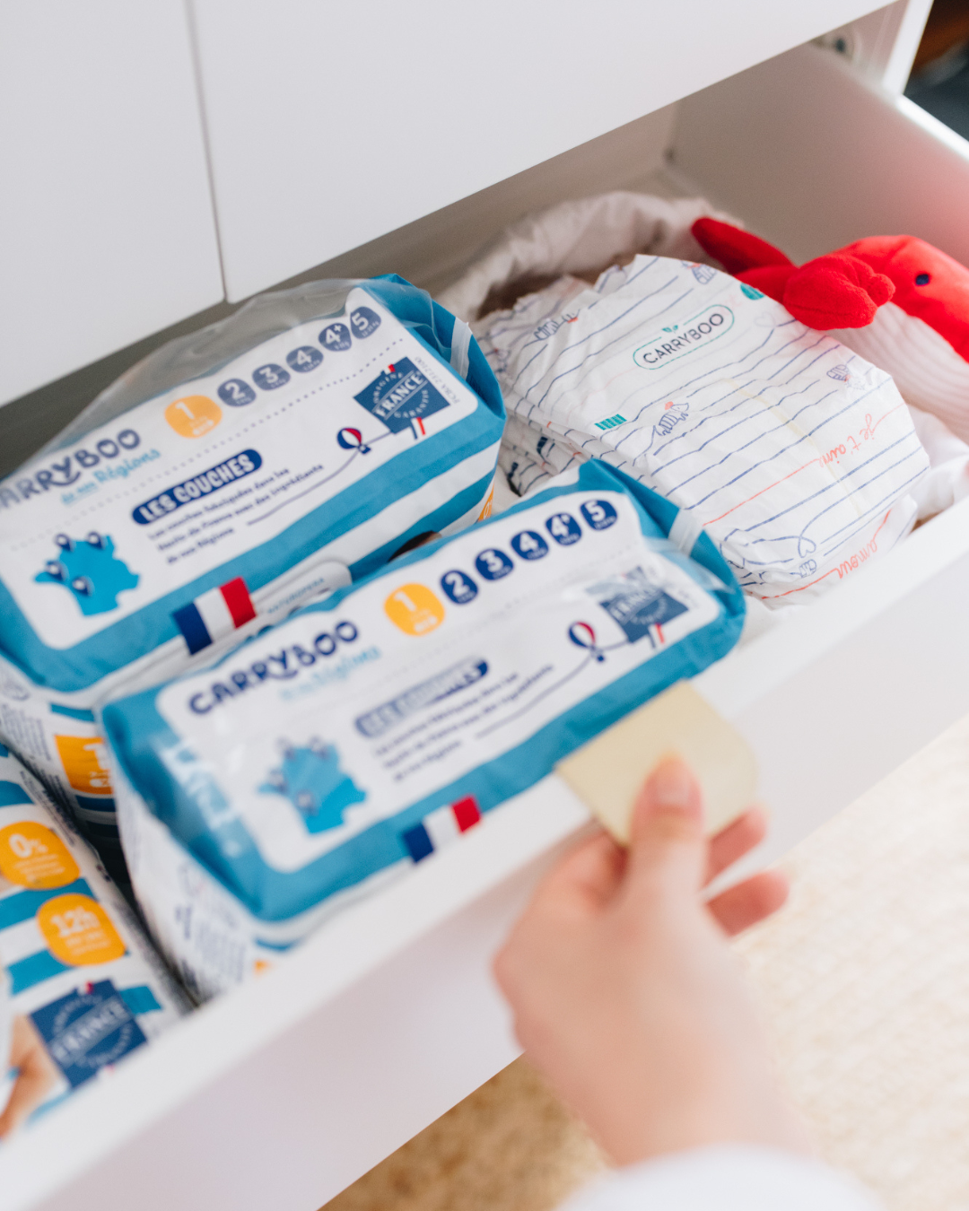 Main qui ouvre un placard de rangement où on aperçoit des paquets de couches taille 1 Carryboo de nos Régions, ainsi que des petites couches Carryboo de nos Régions dans un pochon 