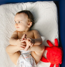Bébé assis, regard sur le côté et souriant sur une table à langer, en couche Carryboo à côté d'un doudou crabe rouge et se tenant les mains et les pieds