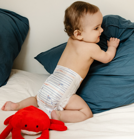 Bébé de profil qui rigole en tenant un oreiller bleu, à côté d'un doudou crabe rouge