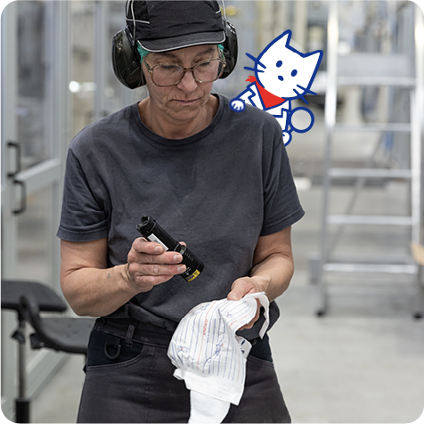 Employée inspectant un produit Carryboo dans une usine, portant un casque et des lunettes de protection, avec un dessin de chat en arrière-plan